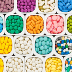 Close-up of multiple square white containers filled with colorful pills and capsules in various shapes and colors, arranged in a neat grid pattern.